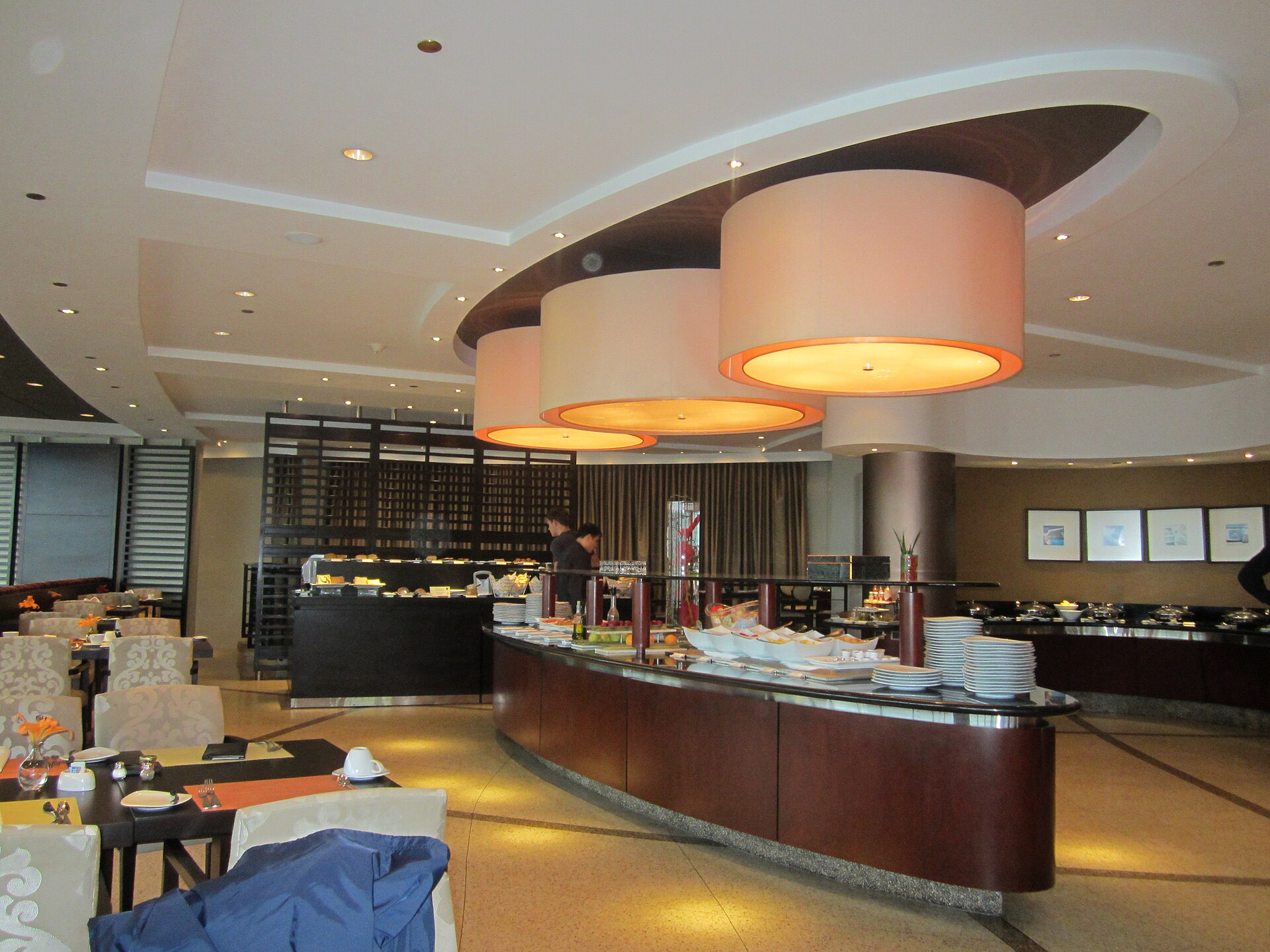 A hotel breakfast buffet: a long serving counter with plates stacked ready, dining tables and pendant lights in the room beyond