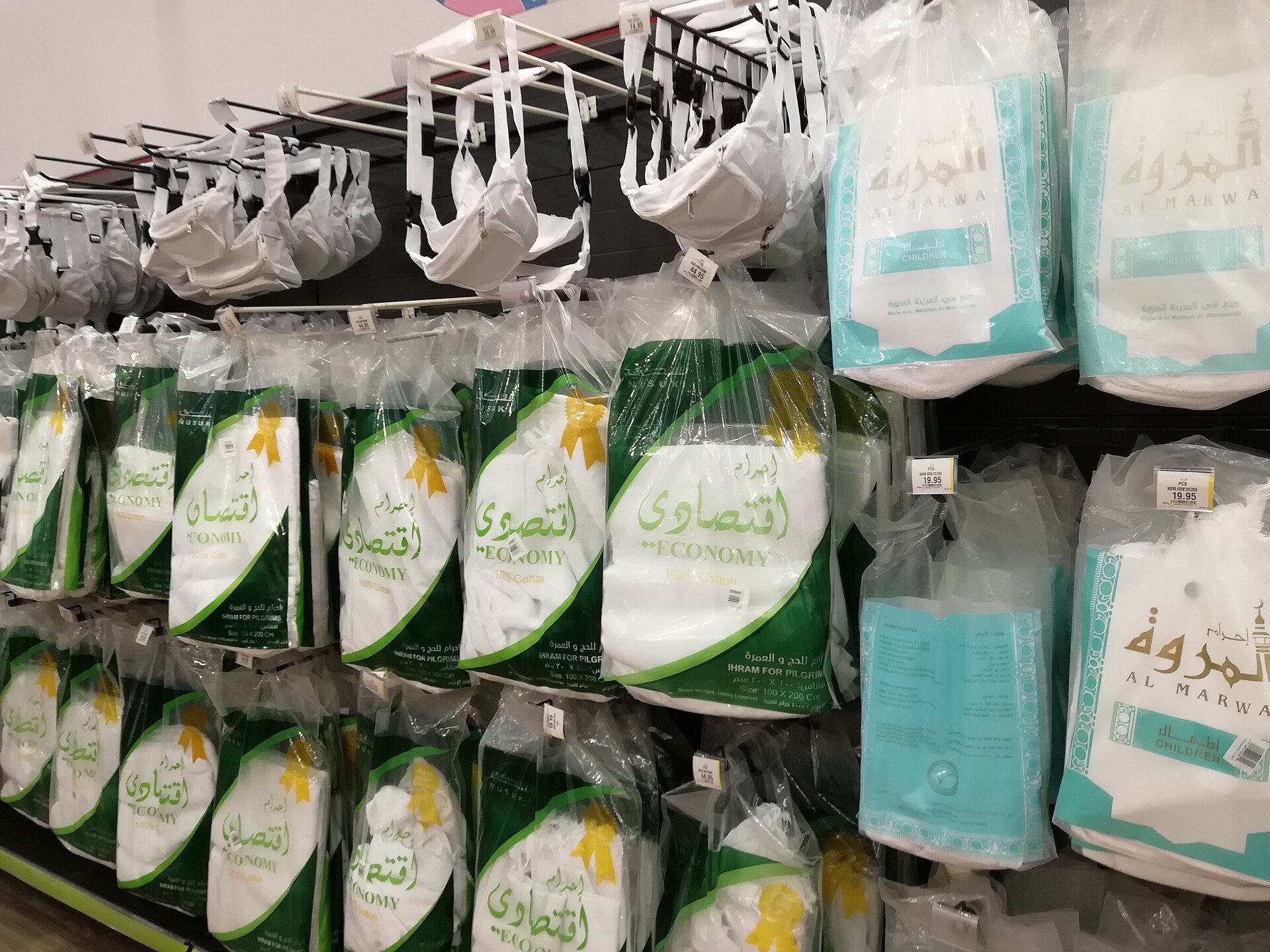 Packaged ihram sets hanging and stacked on a retail rack, white cotton cloth folded inside clear plastic packs with printed labels and price tags