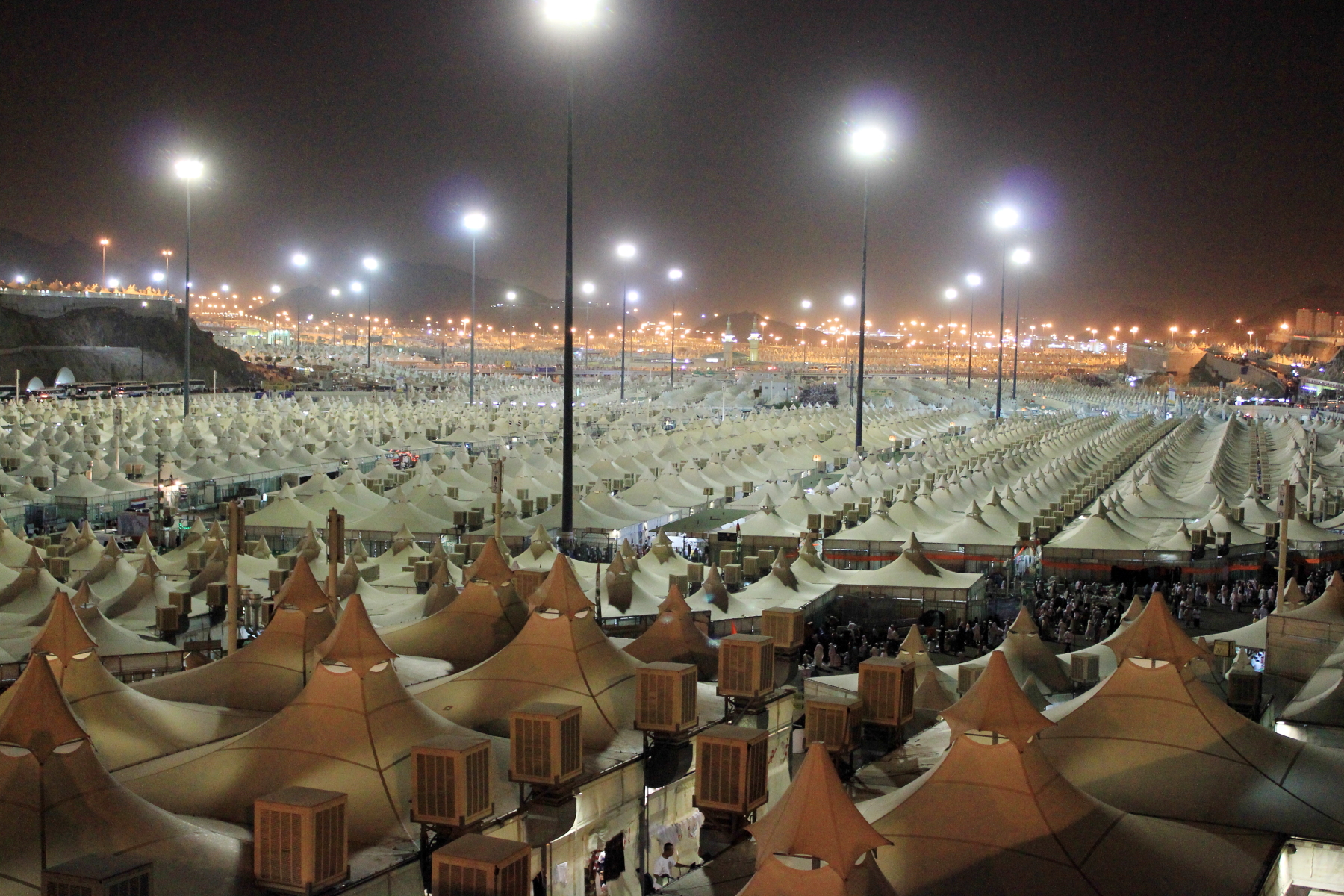 The Mina tent camp at night: rows of white pilgrim tents under floodlights, air-conditioning units between them, rocky hills behind